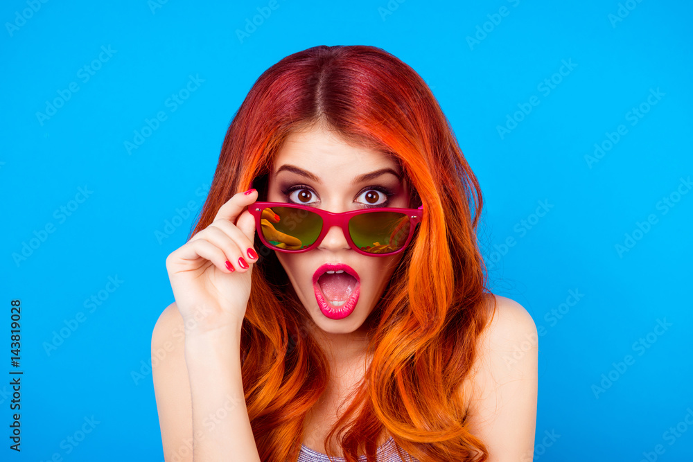 Surprised shocked girl with beautiful red curly long hair holding sunglasses and open mouth while standing on blue background Surprised shocked girl with beautiful red curly long hair holding sunglasses and open mouth while standing on blue background