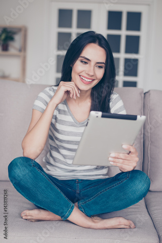 Young pretty pretty woman barefoot in casual clothes (striped shirt and jeans) looks at the tablet while sitting at home on the couch