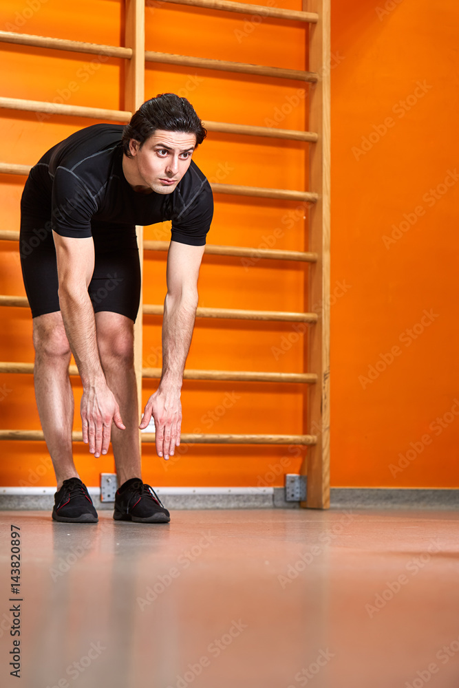 Strong man in the black sportwear stretching arms before gym workout against bright orange wall.