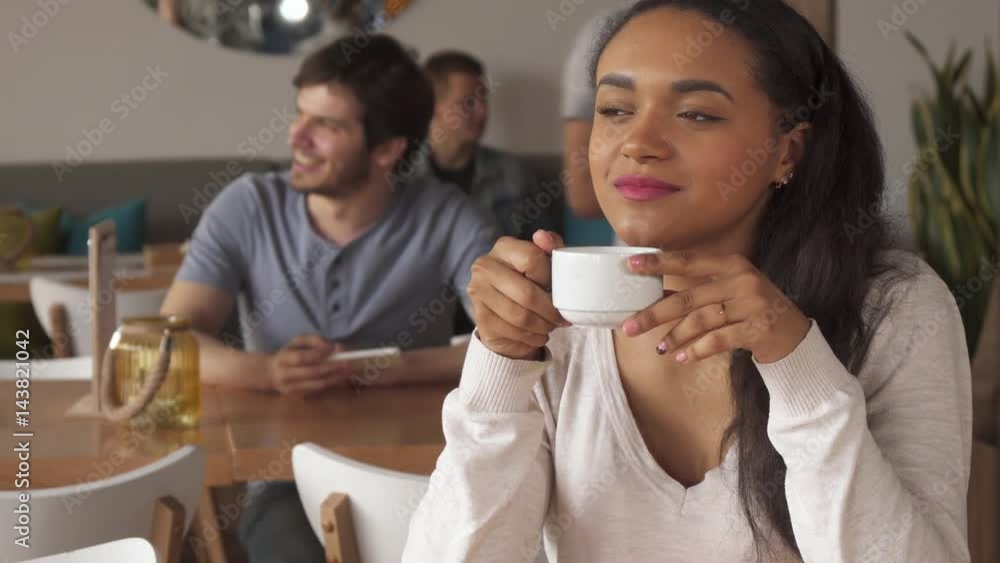 Pretty african american girl dreaming over the cup of coffee at the cafe. Young brown haired woman sitting against background of bearded guy using his smartphone. Close up of attractive lady waiting Pretty african american girl dreaming over the cup of coffee at the cafe. Young brown haired woman sitting against background of bearded guy using his smartphone. Close up of attractive lady waiting