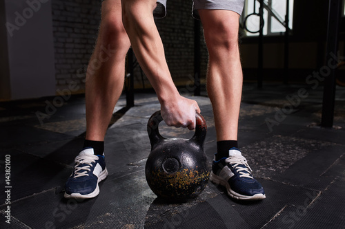 Close-up photo of man's legs and arm while holding kettlebell on the gym floor against dark background.