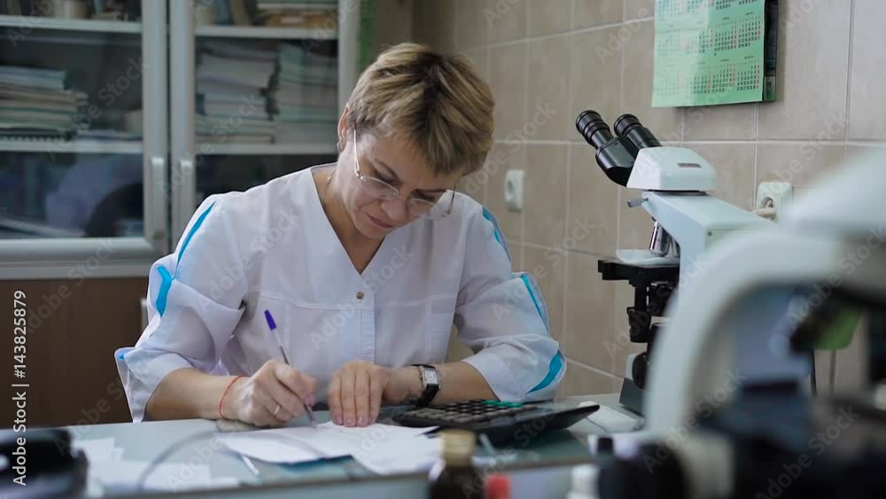The doctor in his office fills in the prescription or medical history. A middle-aged woman with glasses, paper around her, a shelf and a microscope. The doctor in his office fills in the prescription or medical history. A middle-aged woman with glasses, paper around her, a shelf and a microscope.