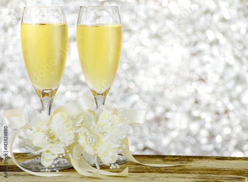 A pair of glasses filled with sparkling bubbly champagne and decorated with ivory colored satin ribbons and silk flowers on a wooden table in front of a sparkly, glittering silver bokeh background