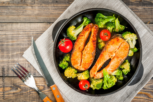 Healthy eating, diet. Baked grilled trout (salmon) with vegetable garnish - broccoli, tomatoes. In a portioned frying pan, on a wooden table. Top view copy space
