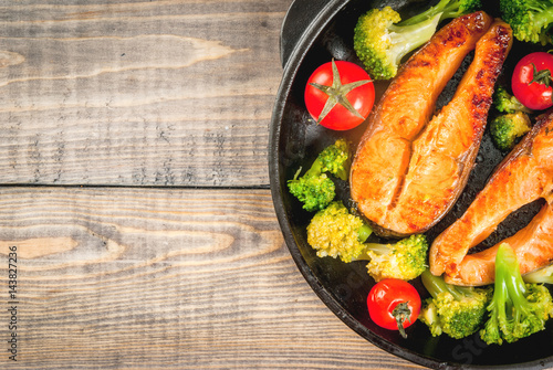 Healthy eating, diet. Baked grilled trout (salmon) with vegetable garnish - broccoli, tomatoes. In a portioned frying pan, on a wooden table. Top view copy space