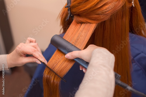 Hairdresser makes hairstyle girl with long red hair in a beauty salon. Straightening hair ironing. Professional hair care.