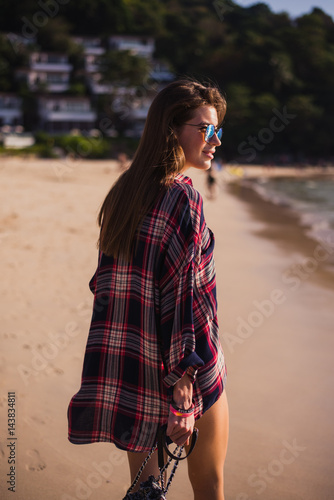 Summer lifestyle portrait of young pretty girl with sportive tanned body, walking to the beach of tropical island. Wearing checkered t-shirt and stylish sunglasses. With small beach bag