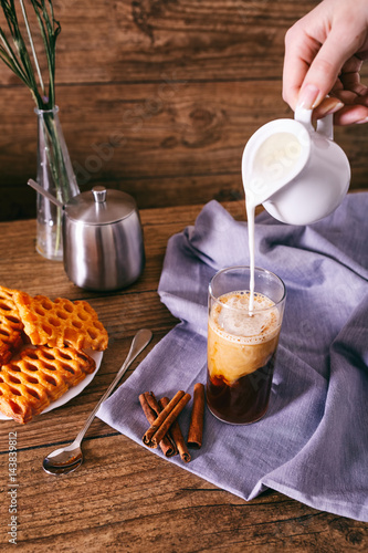 Women's hand with creamer pouring milk in glass with coffee. Cinnamon sticks, homemade cookies and bunch of wildflowers on a wooden background. Breakfast concept.