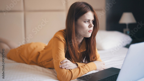a woman in a yellow dress lies on a bed, an open laptop, a puzzled look