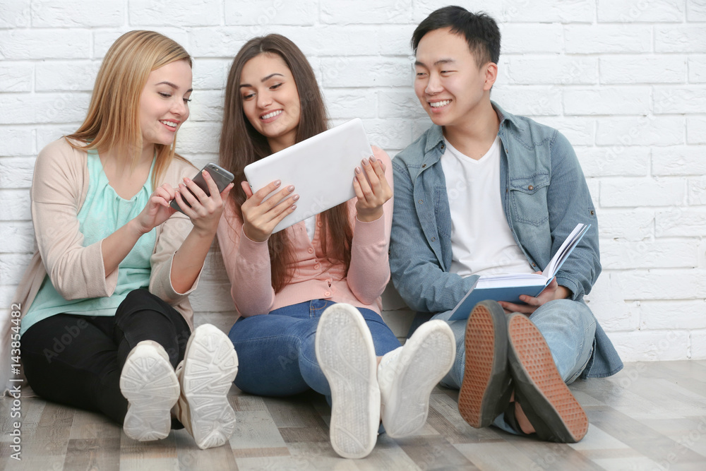 People with book and gadgets sitting on floor near brick wall People with book and gadgets sitting on floor near brick wall