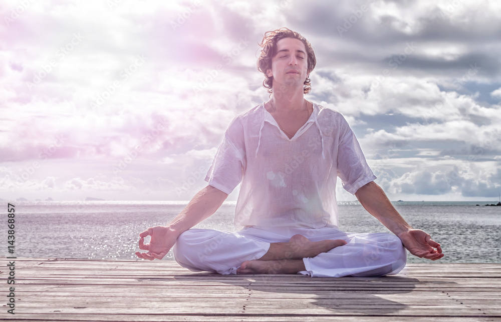 Young man meditating with closed eyes on the beach, clouds in the background