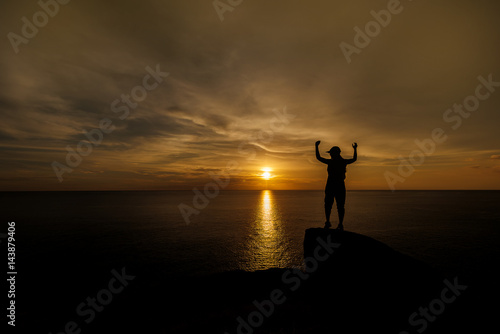 silhouette of woman hand up and standing on rock in sunset. 