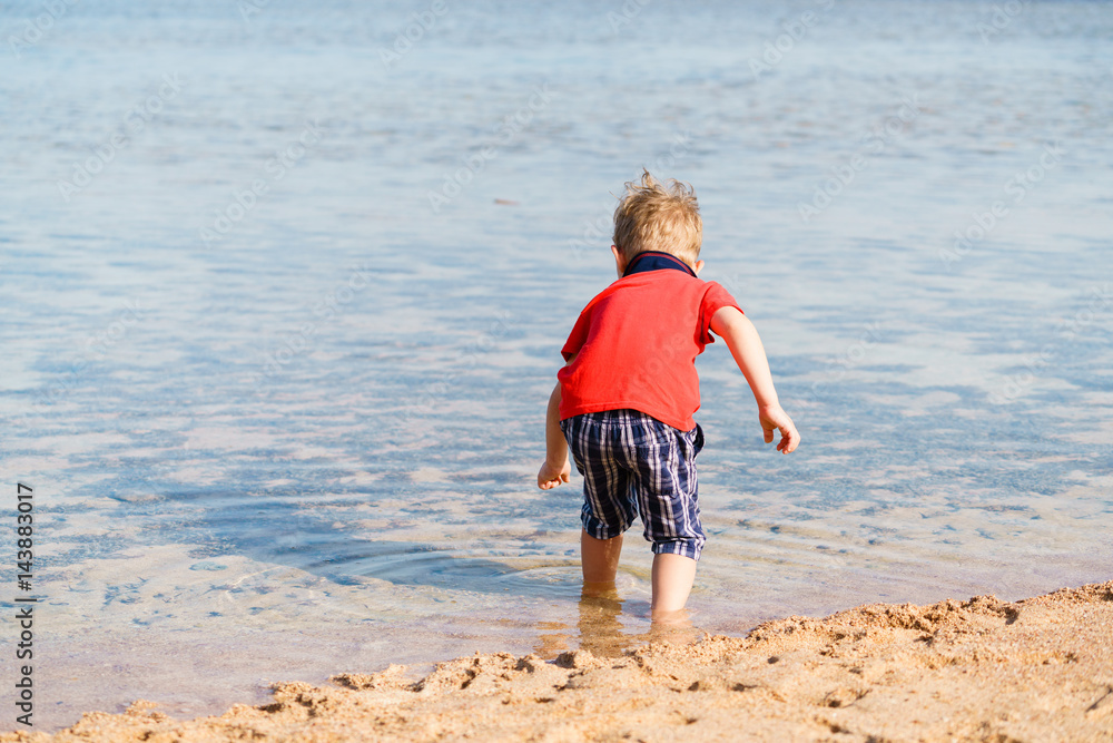 Little cute boy in red polo shirt and checkered shorts is walking barefoot along the sandy beach of the calm sea. Bright sunny summer day.