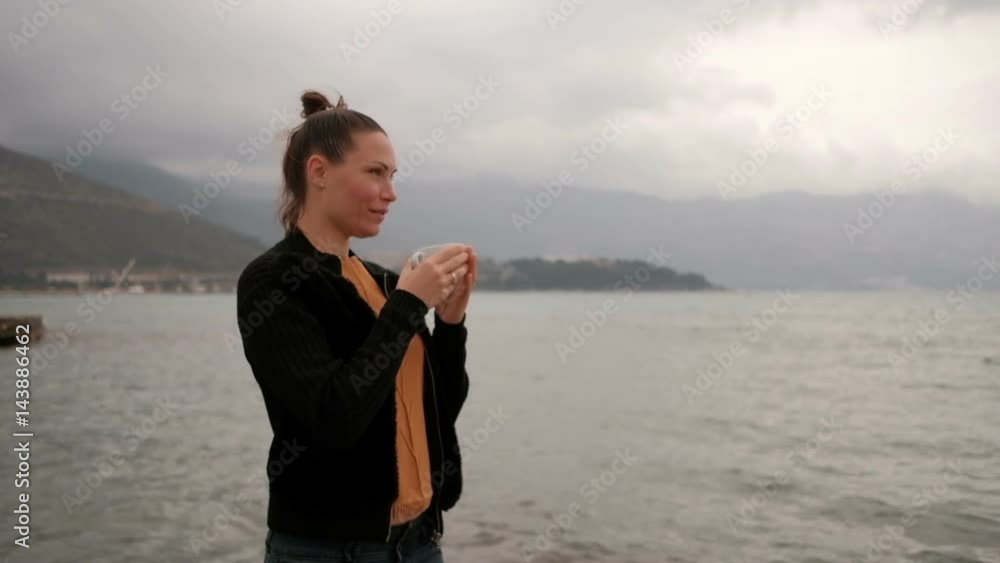 Woman drink coffee standing on lake coast, camera go clockwise. Adult Caucasian female with long hair in black knitted sweater, grey jeans, orange top with pattern of branch under jacket hold white Woman drink coffee standing on lake coast, camera go clockwise. Adult Caucasian female with long hair in black knitted sweater, grey jeans, orange top with pattern of branch under jacket hold white