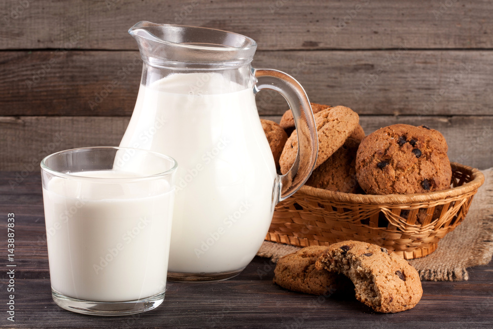 jug and glass of milk with oatmeal cookies in a wicker basket on a wooden background