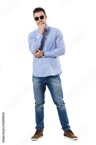 Young smart casual businessman thinking with hands on chin wearing sunglasses. Full body length portrait isolated over white background.