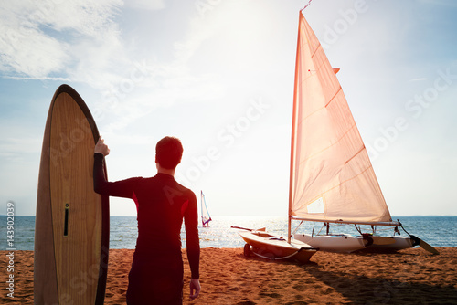 Fotografia Surfer man standing on the beach looking out to the sea with sky background and windsurfing in the wind through the waves at the sea