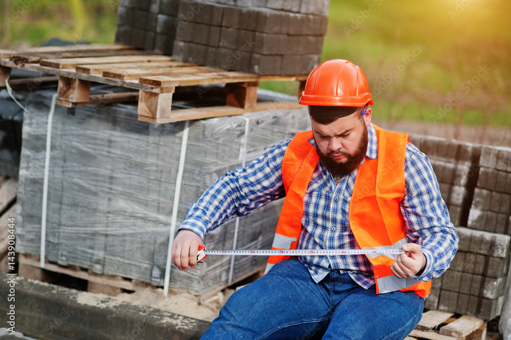 Brutal beard worker man suit construction worker in safety orange helmet against pavement with a tape measure in hand.