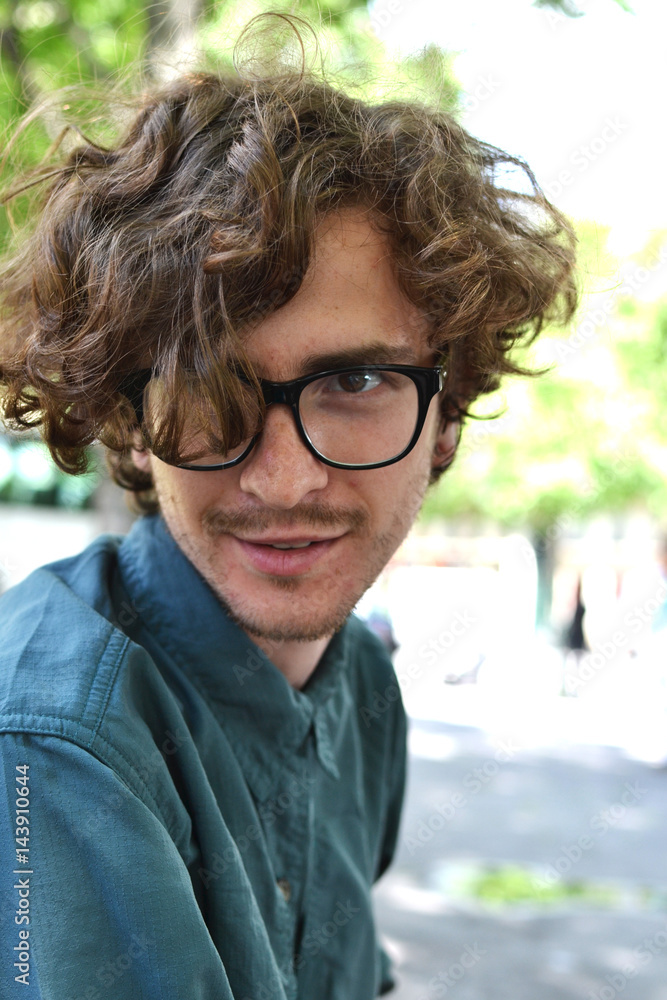 Beautiful young curly-haired guy in black-rimmed glasses travel in Europe on the background of the attractions in the Park, waiting for a date with a girl, resting, summer, spring Stock Photo | Beautiful young curly-haired guy in black-rimmed glasses travel in Europe on the background of the attractions in the Park, waiting for a date with a girl, resting, summer, spring Stock Photo |