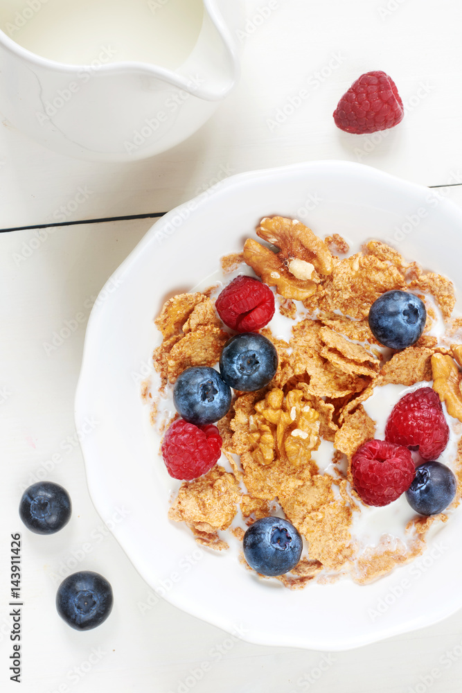 Vertical top view of bowl of cornflakes with raspberry, blueberry, walnut and fresh milk on white wooden table. 