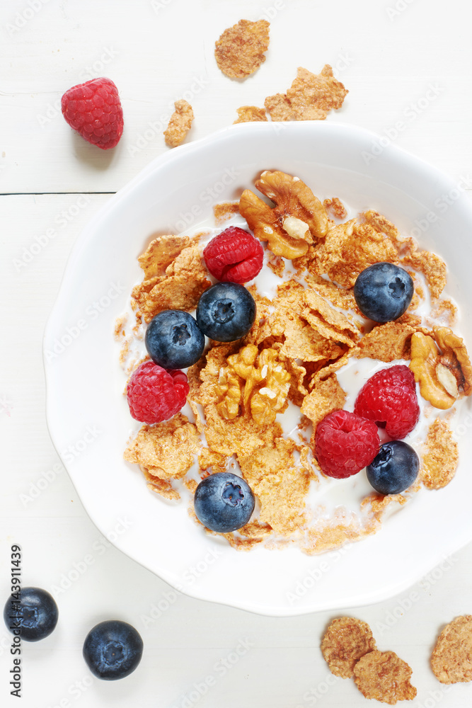 Vertical top view of bowl of cornflakes with milk, raspberry, blueberry and walnut on white wooden table. Vertical top view of bowl of cornflakes with milk, raspberry, blueberry and walnut on white wooden table.
