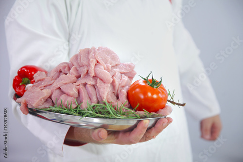 A chef is holding a tray with raw meat in his hands, a tomato and a red pepper. It is going to put it into the oven.