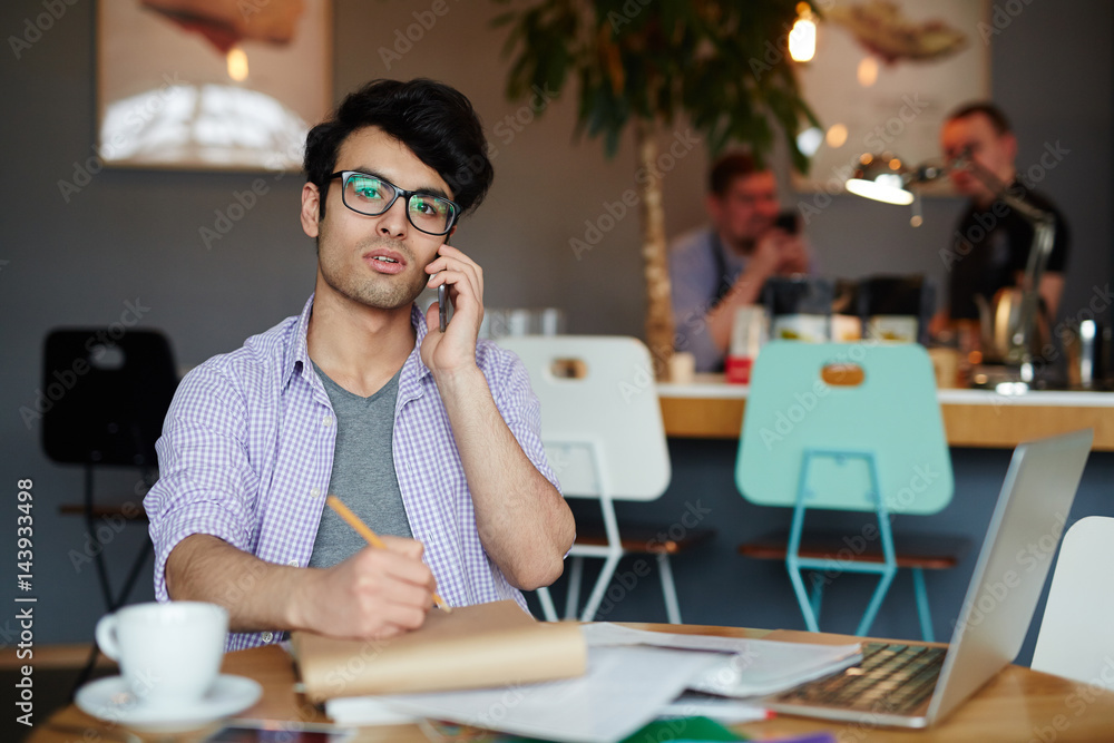 Portrait of young creative man wearing casual clothes and glasses  making notes while calling by phone and working with laptop in cafe