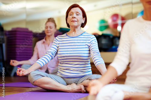 Mature barefoot woman sitting in pose of lotus during meditation in gym