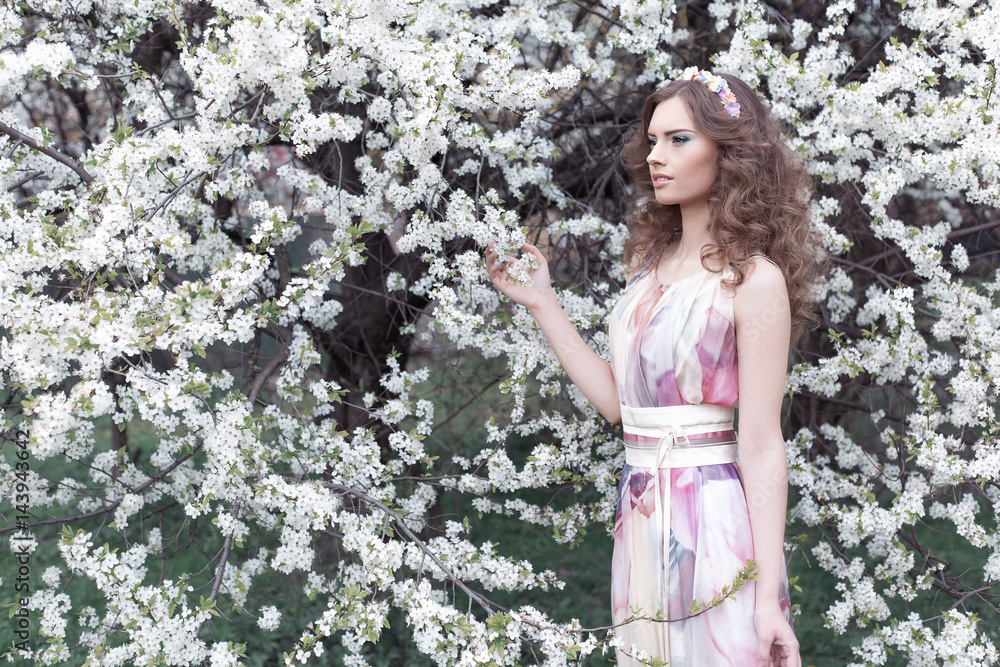 Beautiful young girl with curls in an air colored dress with a wreath on her head walking in the park near a cherry blossom on a sunny day Beautiful young girl with curls in an air colored dress with a wreath on her head walking in the park near a cherry blossom on a sunny day