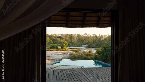 A slow linear dolly shot from the inside of a luxurious private African lodge with a view onto the patio deck with swimming pool and river in the distance just after dawn, at sunrise