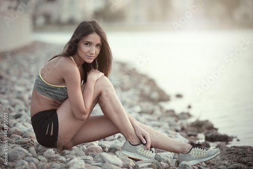 Beautiful athletic fitness model sitting at the edge of a calm lake with a little smile thinking about her next exercise