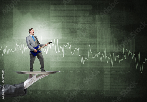 Businessman on metal tray playing electric guitar against wall with charts background