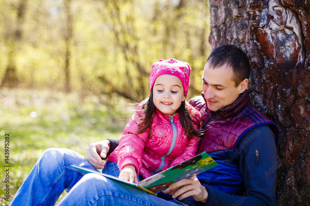 Father reading the book to his young daughter under the tree in the park with his wife. Sitting on the meadow under the tree. Family relaxing outdoors Father reading the book to his young daughter under the tree in the park with his wife. Sitting on the meadow under the tree. Family relaxing outdoors