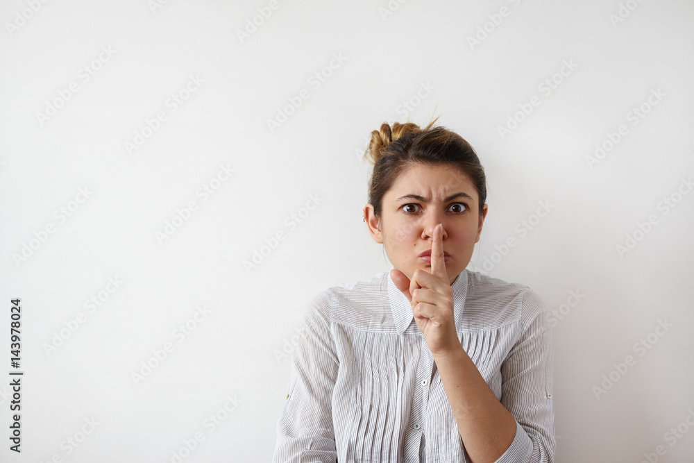 Portrait of funny young Caucasian woman in white shirt with hair bun looking seriously at the camera, holding index finger at lips, asking to keep silence or not tell anyone her secret, saying 'Tsss' Portrait of funny young Caucasian woman in white shirt with hair bun looking seriously at the camera, holding index finger at lips, asking to keep silence or not tell anyone her secret, saying 'Tsss'