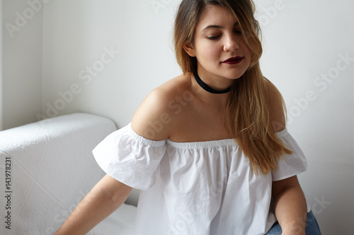 Attractive young woman in white blouse with poen shoulders looking down posing at home interior. Natural light from the window