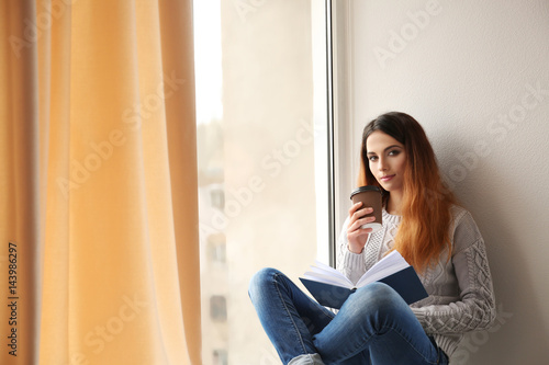 Beautiful young woman drinking coffee and reading book while sitting on window sill at home