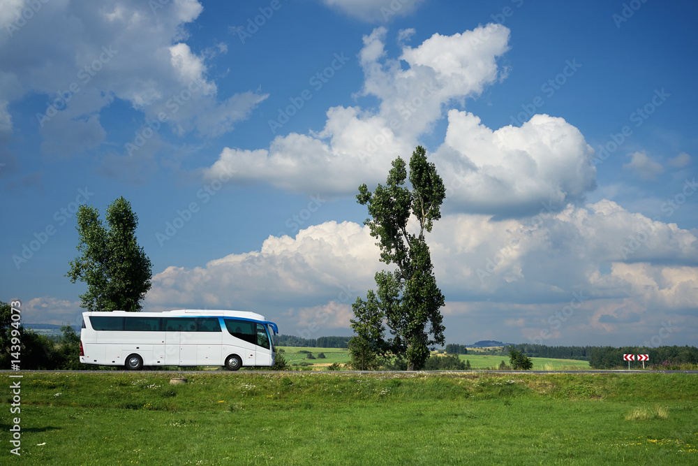 The white bus traveling on the road next to tall trees in a rural landscape under a blue sky with dramatic white clouds