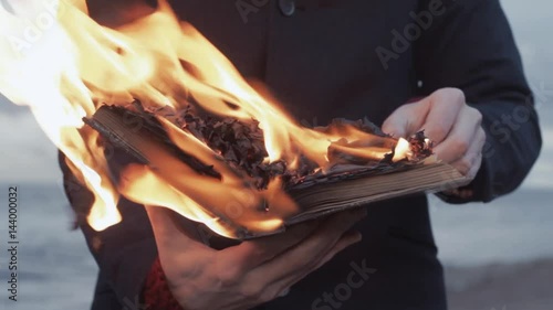 Young man with burning book in hand standing on coast storm clouds on background