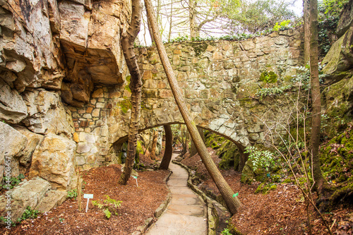 A stone bridge over a pedestrian footpath beside a rock cliff dotted with trees and green foliage