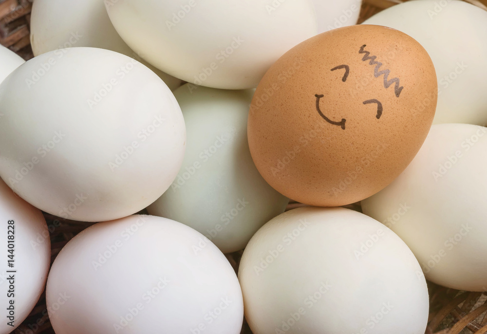 Closeup brown chicken egg with paint in smile face on pile of white duck egg on wood basket background Closeup brown chicken egg with paint in smile face on pile of white duck egg on wood basket background
