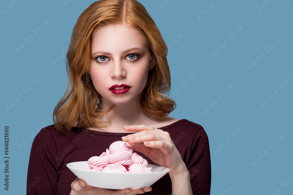 Portrait of a beautiful happy young blonde girl in the studio on a on a blue background with a plate with a marshmallow on hand Portrait of a beautiful happy young blonde girl in the studio on a on a blue background with a plate with a marshmallow on hand