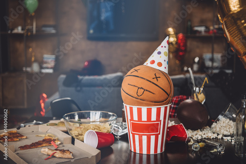 Photography Close-up view of basketball ball with funny face in popcorn box and messy table