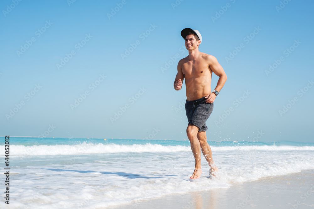 Young sporty man running along beautiful beach, looking happy.