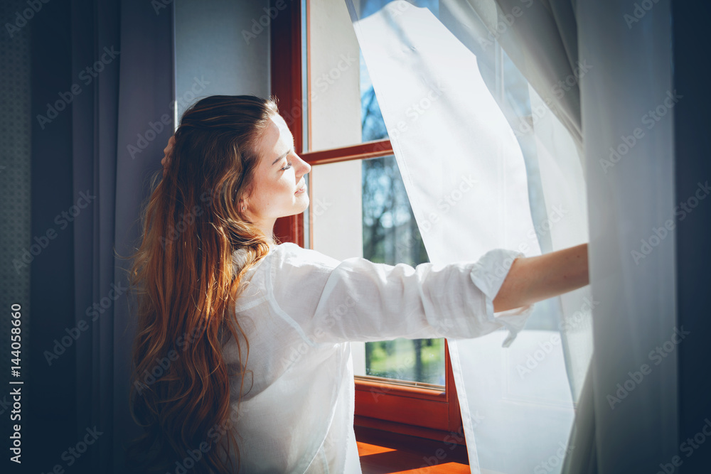 Naklejka premium Pretty woman in modern apartment opening window curtains after wake up