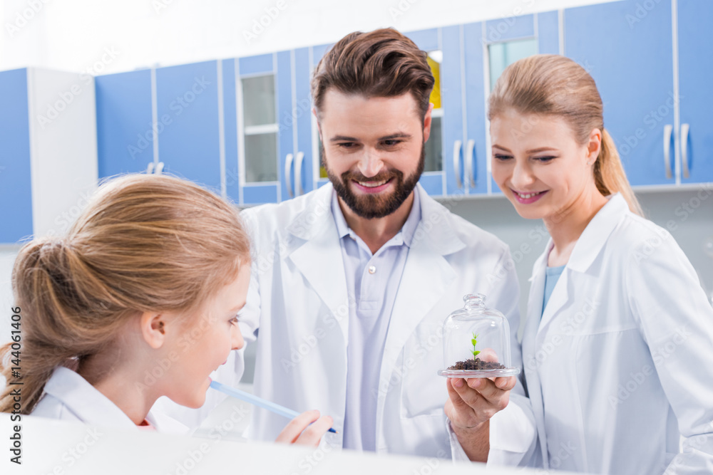 Smiling teachers scientists with green plant in soil looking at little student in laboratory Smiling teachers scientists with green plant in soil looking at little student in laboratory