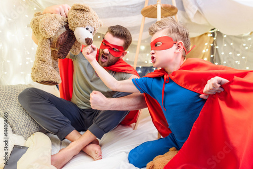 Father and son in superhero costumes playing with teddy bear in blanket fort