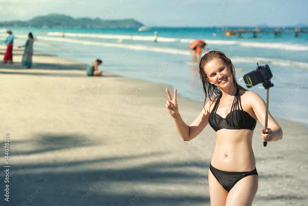 Young woman taking selfie with smartphone. Sexy girl in black bikini on beach taking selfie using monopod