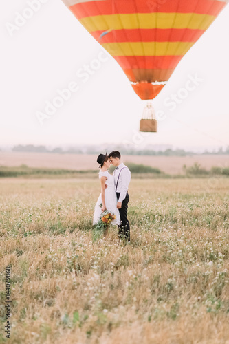 Hugging head-to-head happy newlyweds dressed in the vintage style are standing in the front of the flying airballoon among the spring field.