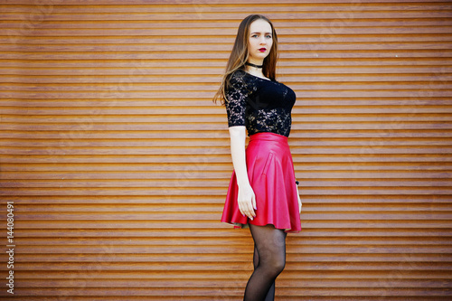 Portrait of girl with black choker necklace on her neck and red leather skirt against orange shutter.