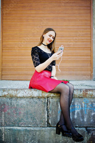 Portrait of girl with black choker on her neck  red leather skirt and mobile phone at hand with headphones against orange shutter.